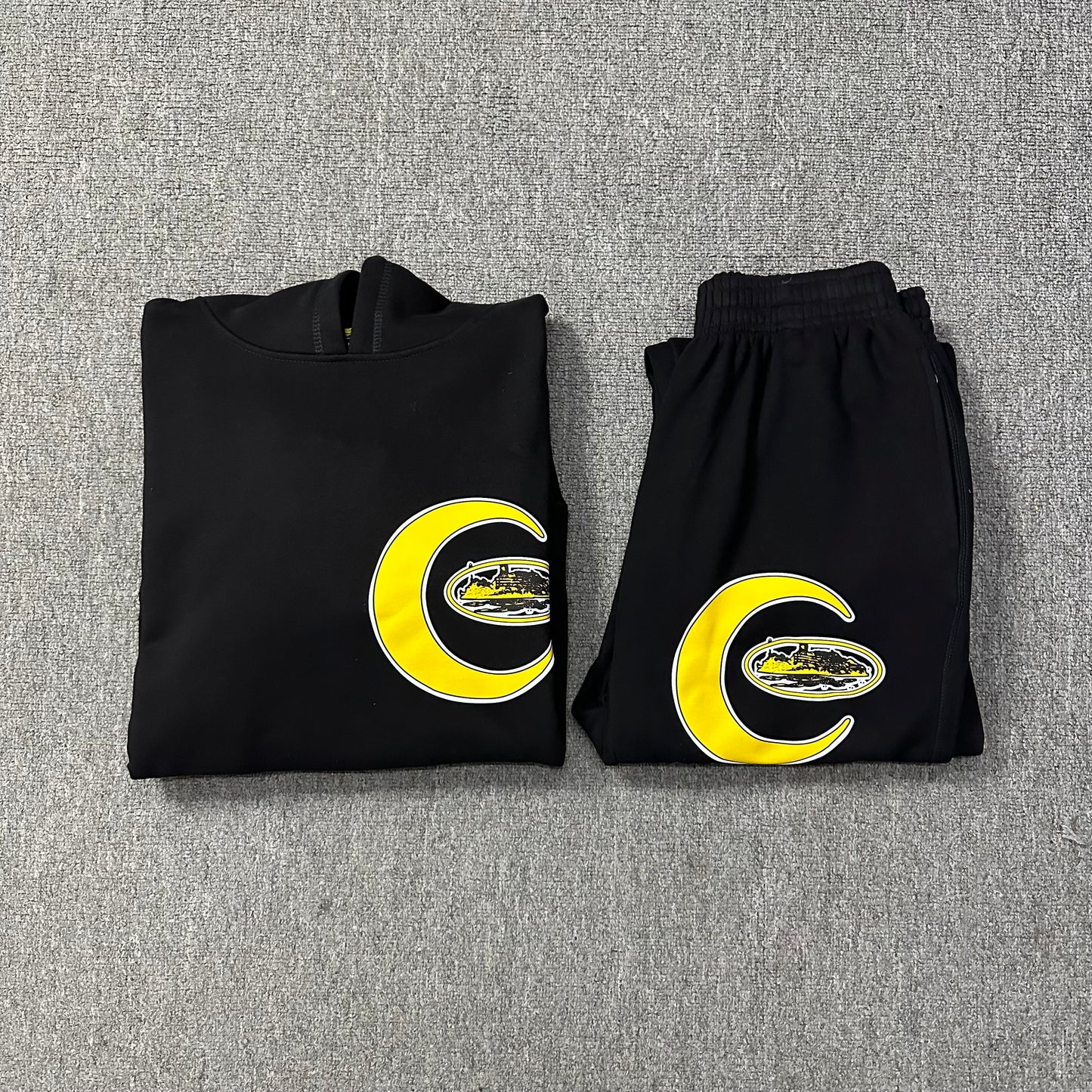 Folded black hoodie and pants with yellow crescent moon and letter 'C' design on a gray textured background