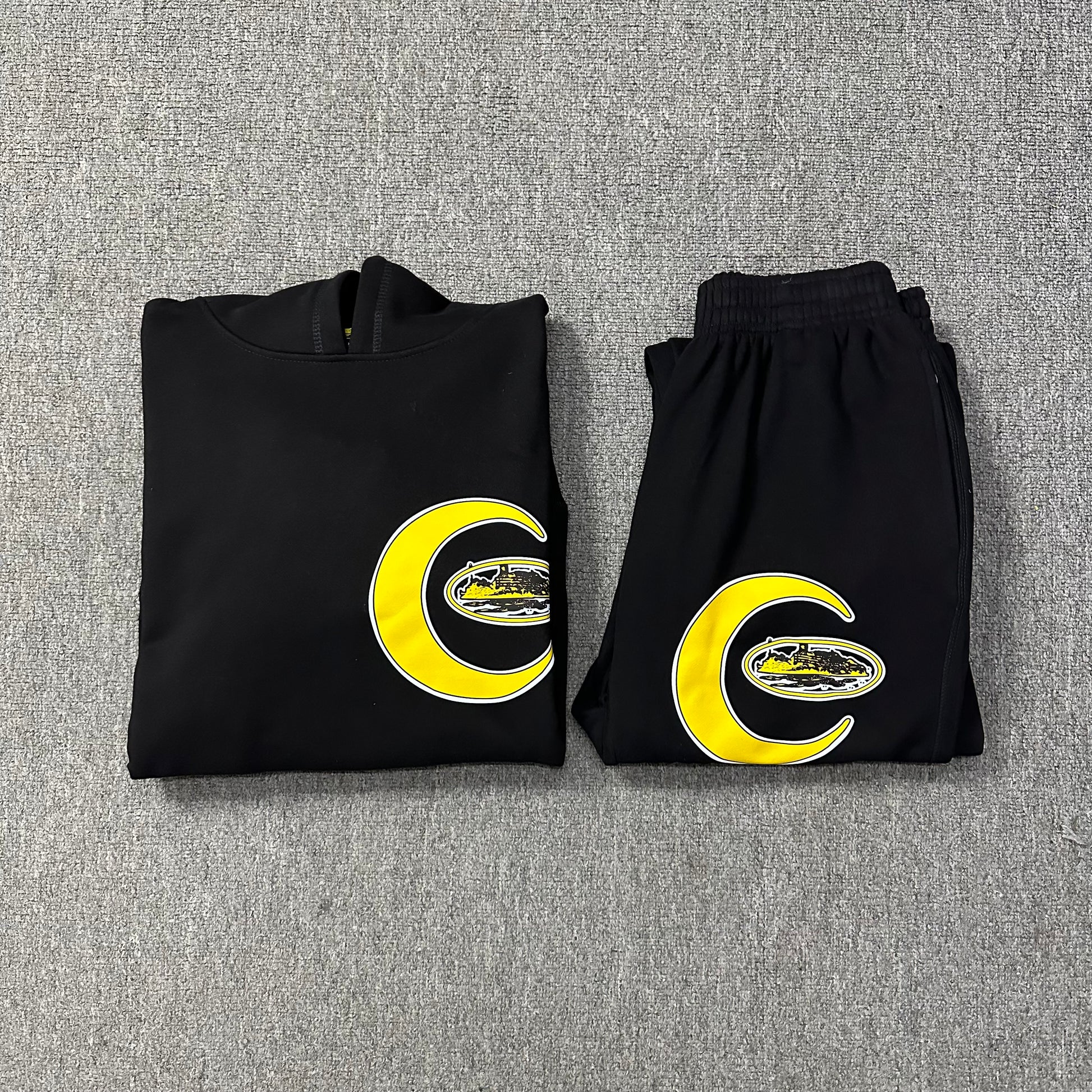 Folded black hoodie and pants with yellow crescent moon and letter 'C' design on a gray textured background
