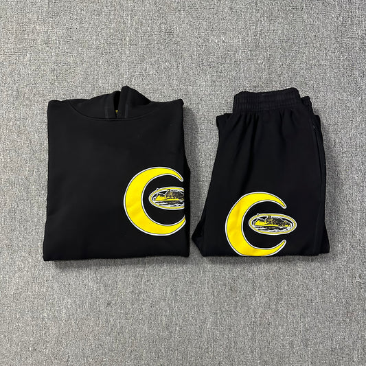 Folded black hoodie and pants with yellow crescent moon and letter 'C' design on a gray textured background
